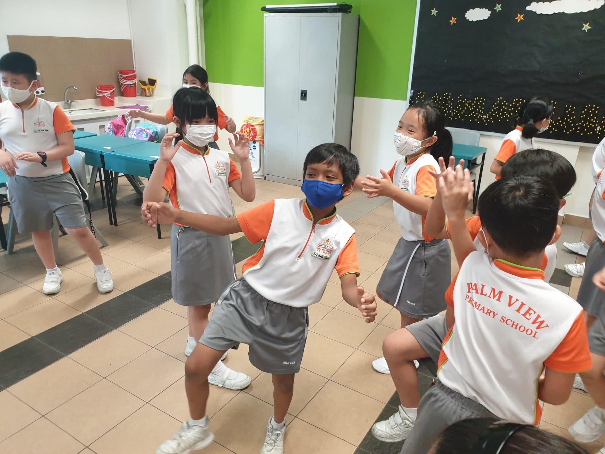 Children in Palm View Primary School uniforms and face masks dance together in a classroom.